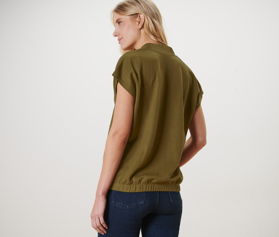 Une femme blonde, vue de dos, porte une blouse verte à encolure en V et un jean.