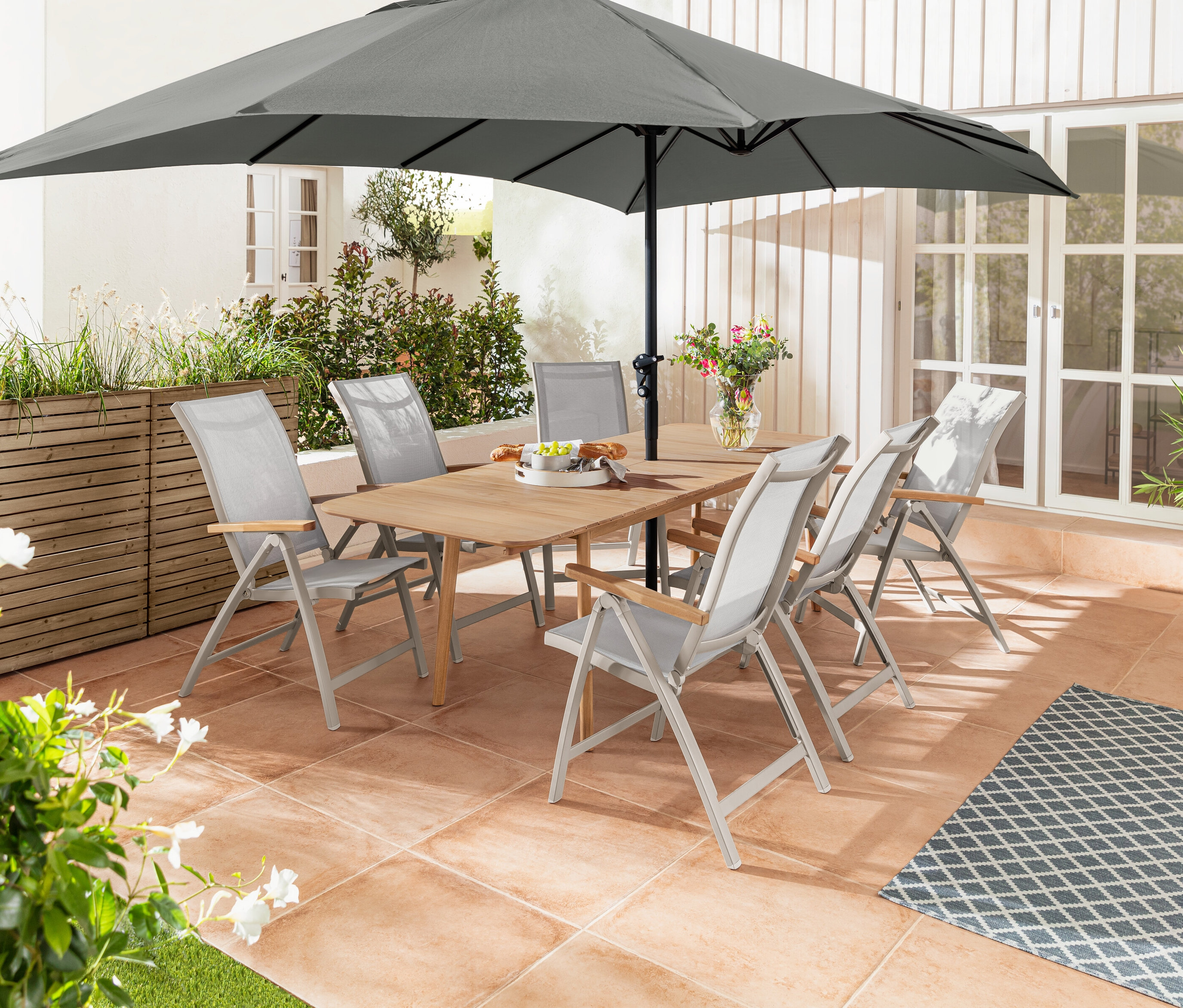 Une table à rallonge en teck est entourée de six chaises « Liska » avec dossier haut réglable. À l'arrière-plan, un séparateur de jardinière « Tinus » avec armoire de rangement.