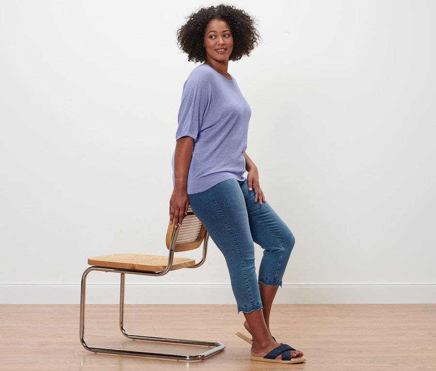 Une femme est assise de côté sur une chaise et porte un T-shirt en maille fine en lin mélangé, un tregging 7/8 à broderie ajourée et des sandales confortables.