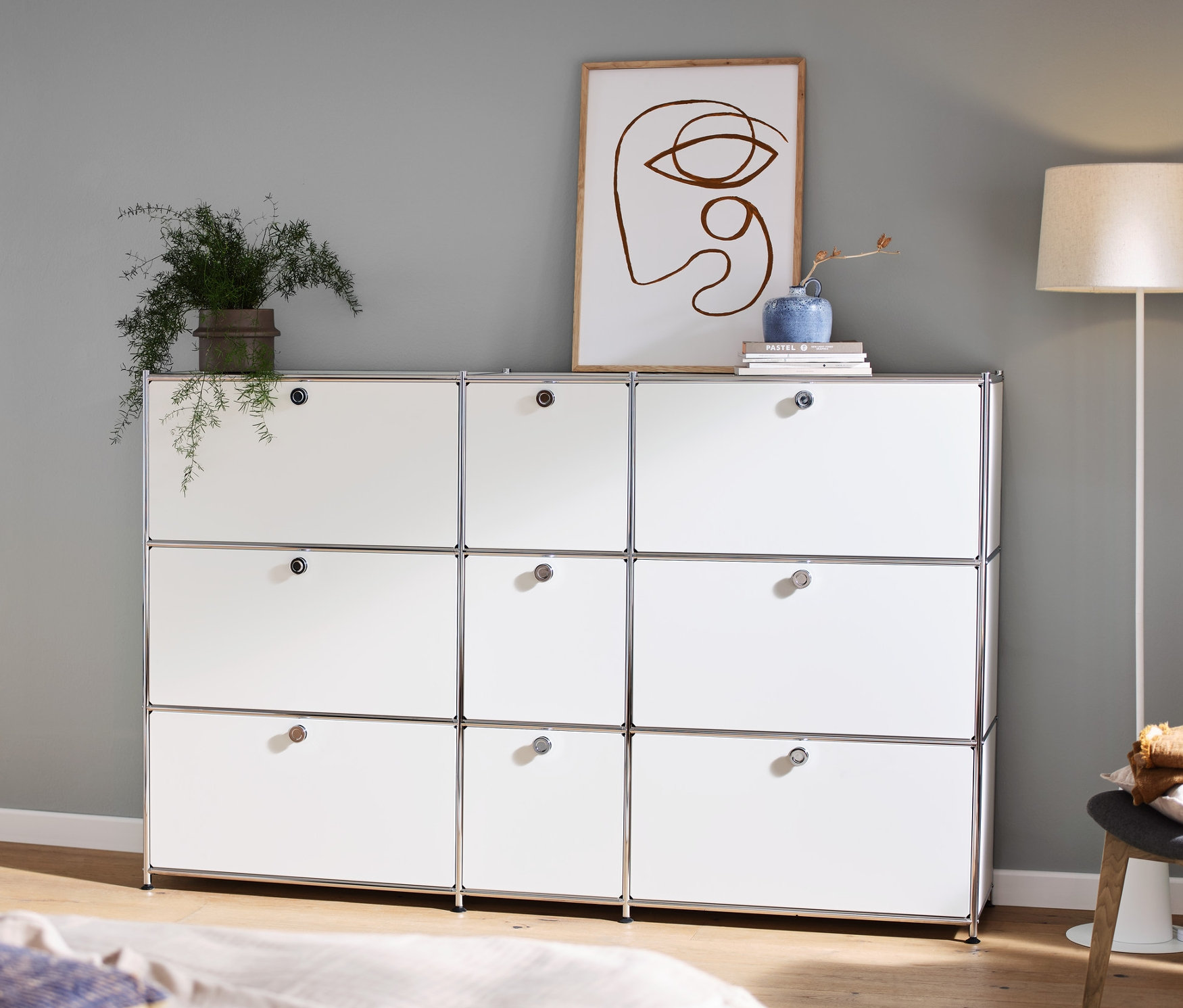 Dans la pièce, il y a une commode « CN3 » blanche avec 9 abattants, sur laquelle se trouve une décoration, à gauche une console et un lampadaire avec abat-jour textile.