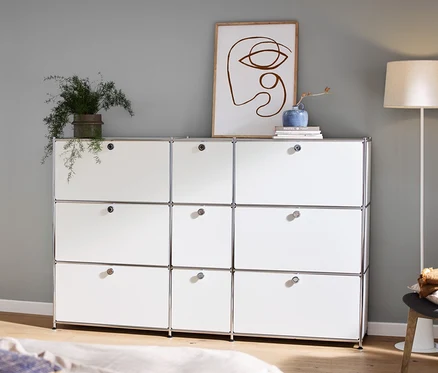 Dans la pièce, il y a une commode « CN3 » blanche avec 9 abattants, sur laquelle se trouve une décoration, à gauche une console et un lampadaire avec abat-jour textile.