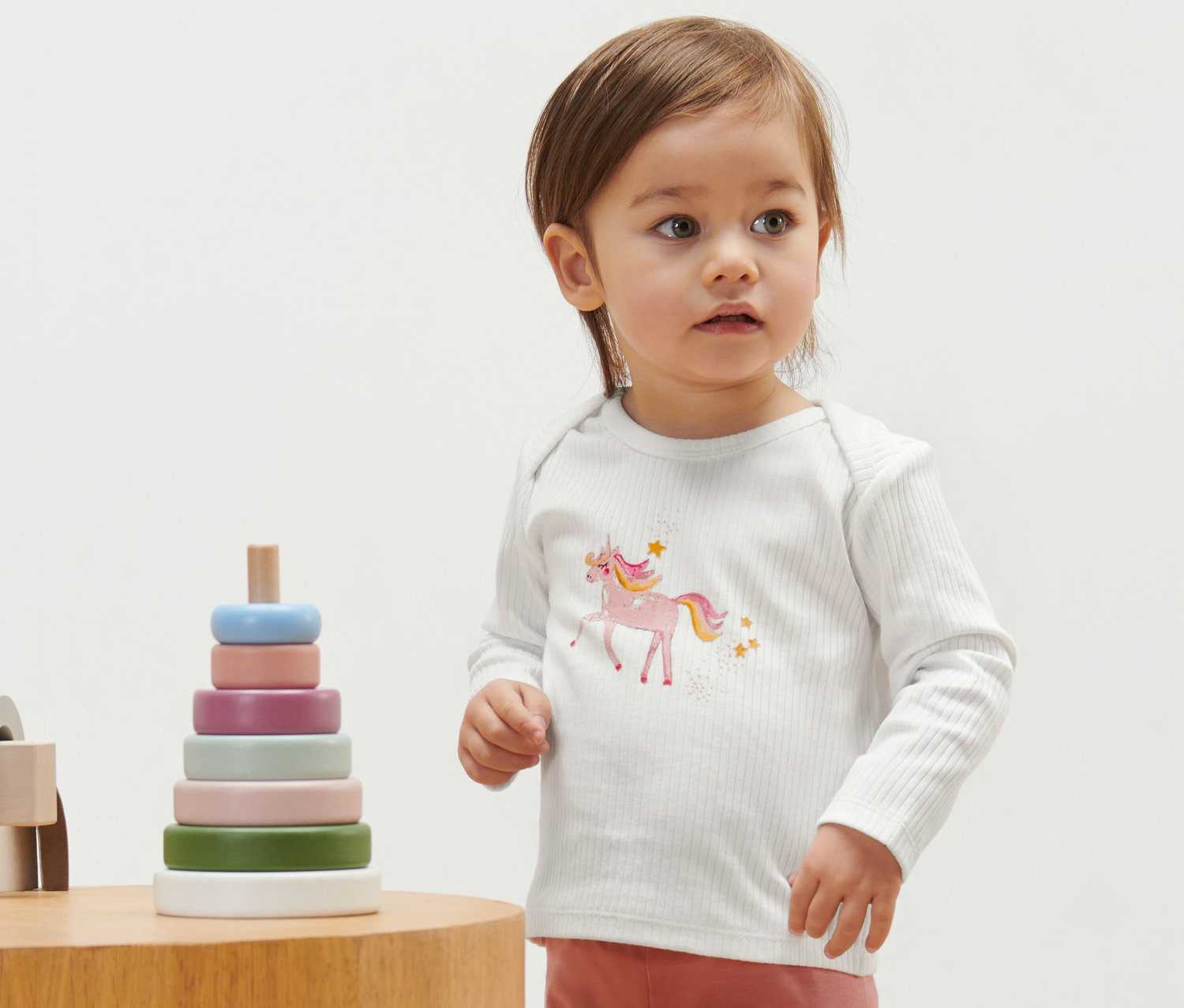 Un bébé porte un t-shirt côtelé avec un motif de rhinocéros. À côté de lui se trouve un jouet en bois.