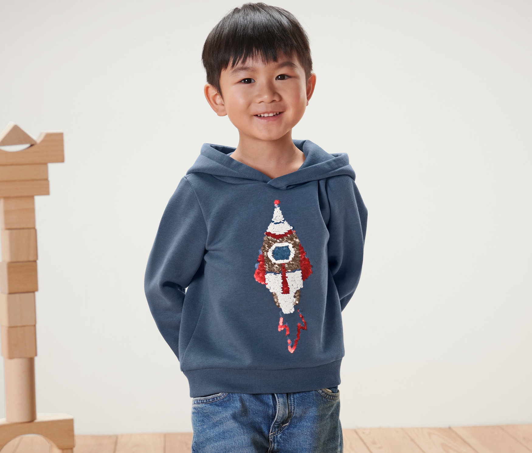 Un garçon souriant pose, portant un sweatshirt pour enfant avec motif en paillettes réversibles, fusée.