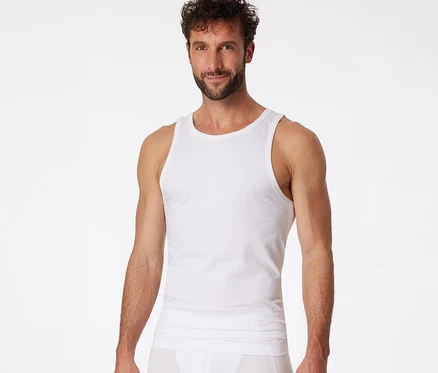 Homme souriant aux cheveux bouclés foncés et à la barbe, portant un maillot de corps SCHIESSER blanc et des sous-vêtements blancs.