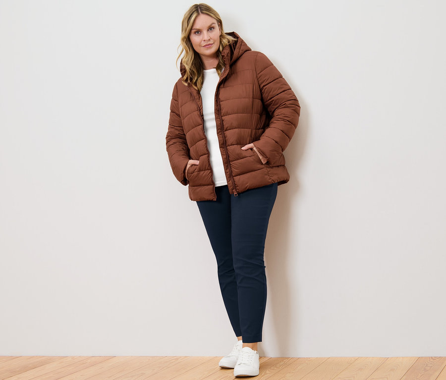 Femme posant en veste matelassée à capuche cognac, pantalon stretch bleu foncé et t-shirt manches 3/4 blanc.