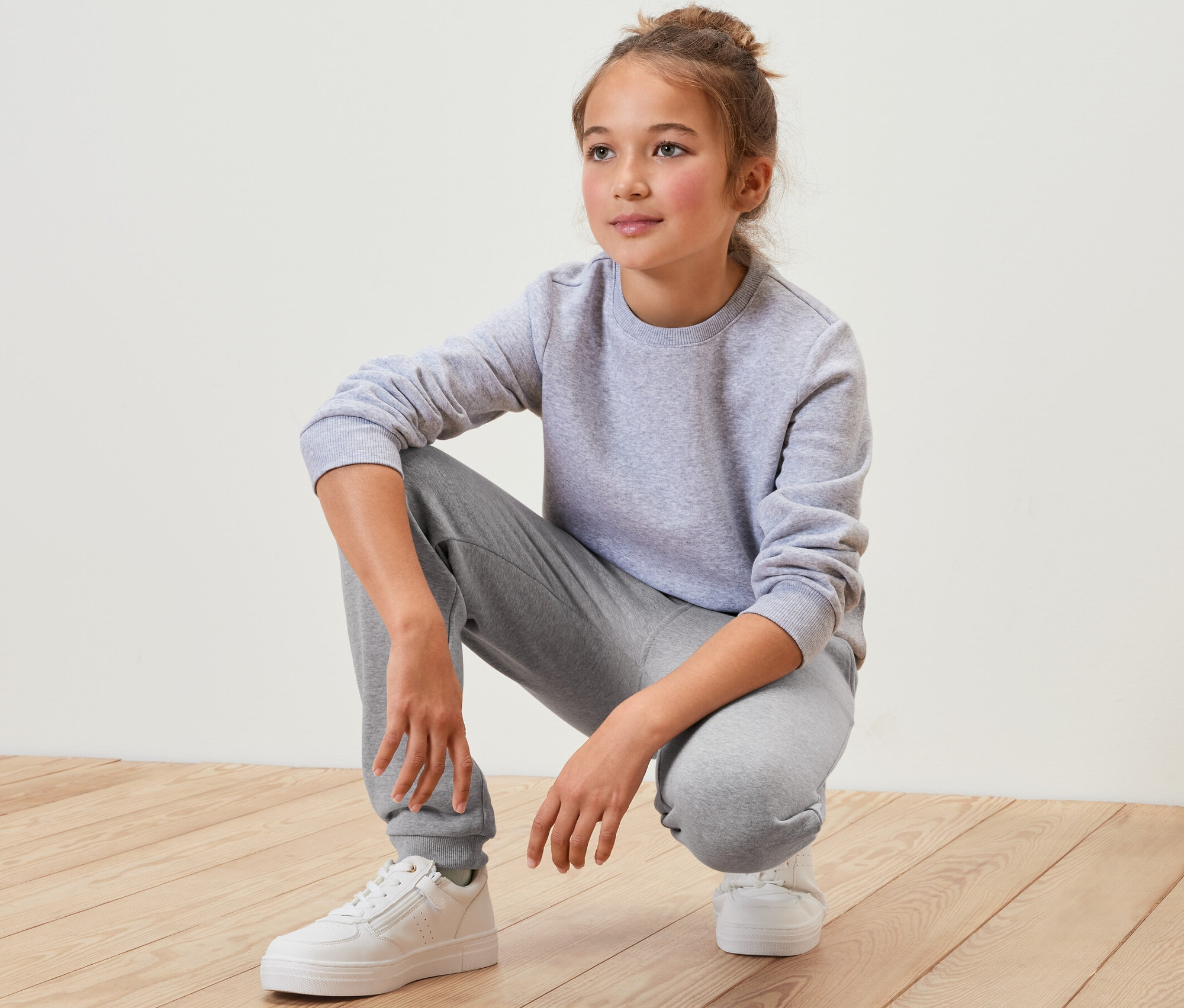 Une fille accroupie sur un plancher en bois, portant un survêtement gris et des chaussures blanches. Elle porte un pantalon en sweat pour enfant, gris.