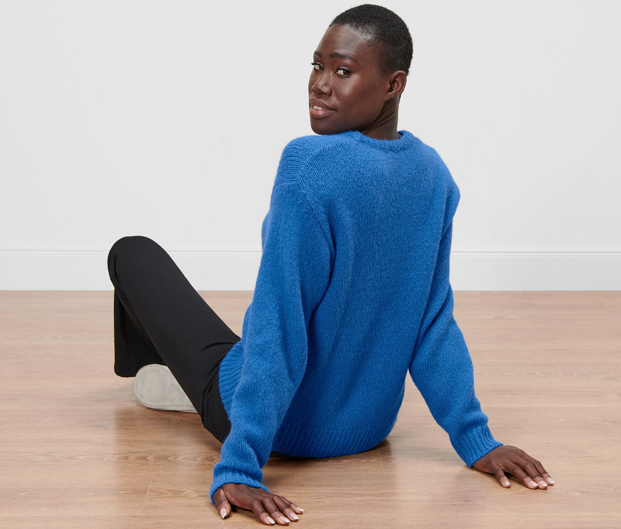 Une femme est assise par terre et regarde par-dessus son épaule, elle porte un pull en grosse maille bleu cobalt avec de la laine.