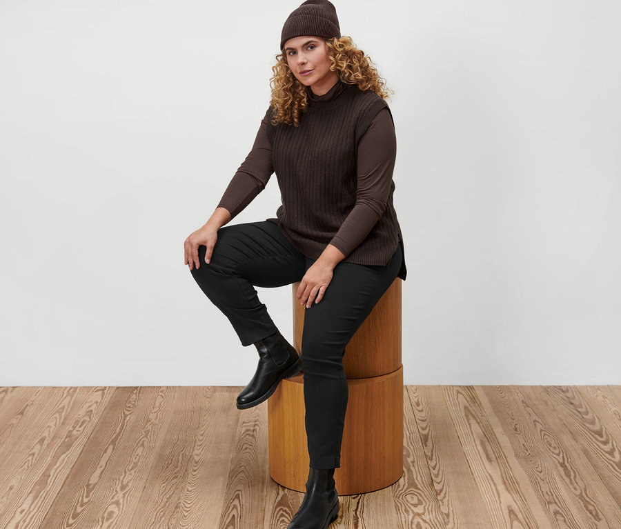 Une femme est assise sur un tabouret en bois et porte un t-shirt col roulé marron, un pull sans manches en maille, un bonnet en maille marron et un pantalon stretch noir.