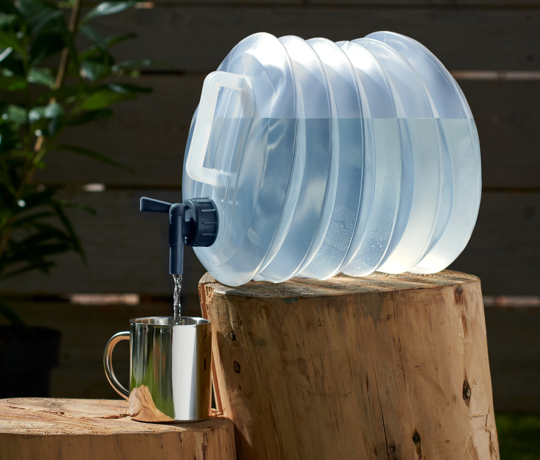 Un jerricane pliable d'environ 10 l est posé sur une bûche et remplit d'eau une tasse argentée.