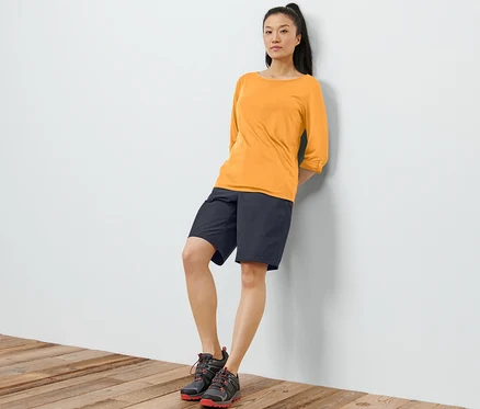 Une femme appuyée contre un mur porte un t-shirt fonctionnel orange à manches 3/4, un short noir et des chaussures.