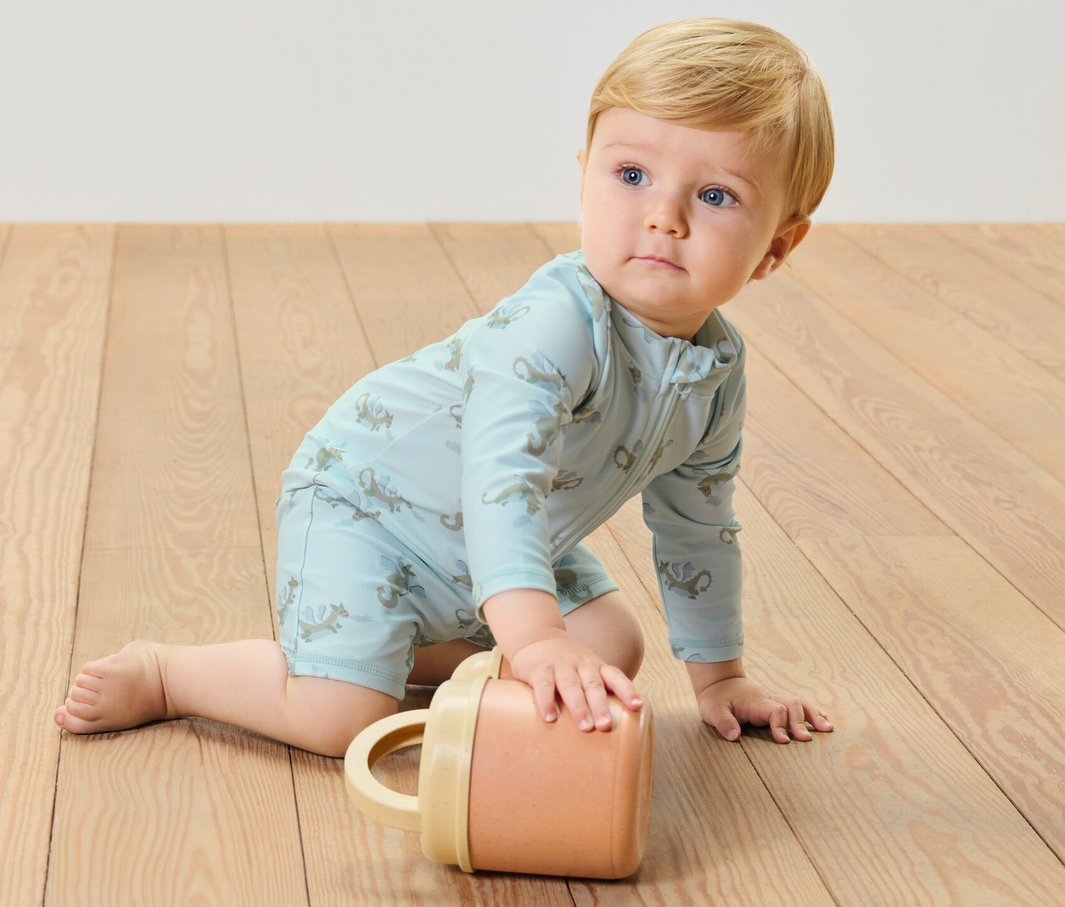 Un bébé rampe sur un plancher en bois, portant une combinaison bleue pour bébé avec un indice de protection UV 80, dragon, tout en jouant avec une tasse.