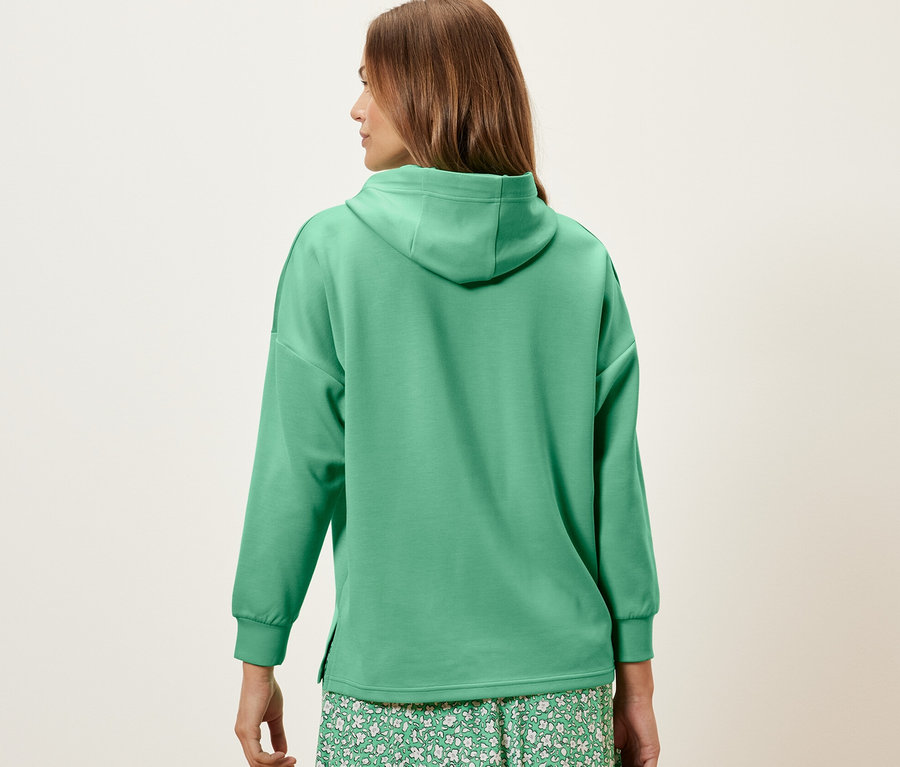 Une femme vue de dos porte un pull à capuche vert en mélange de modal et une jupe à fleurs.