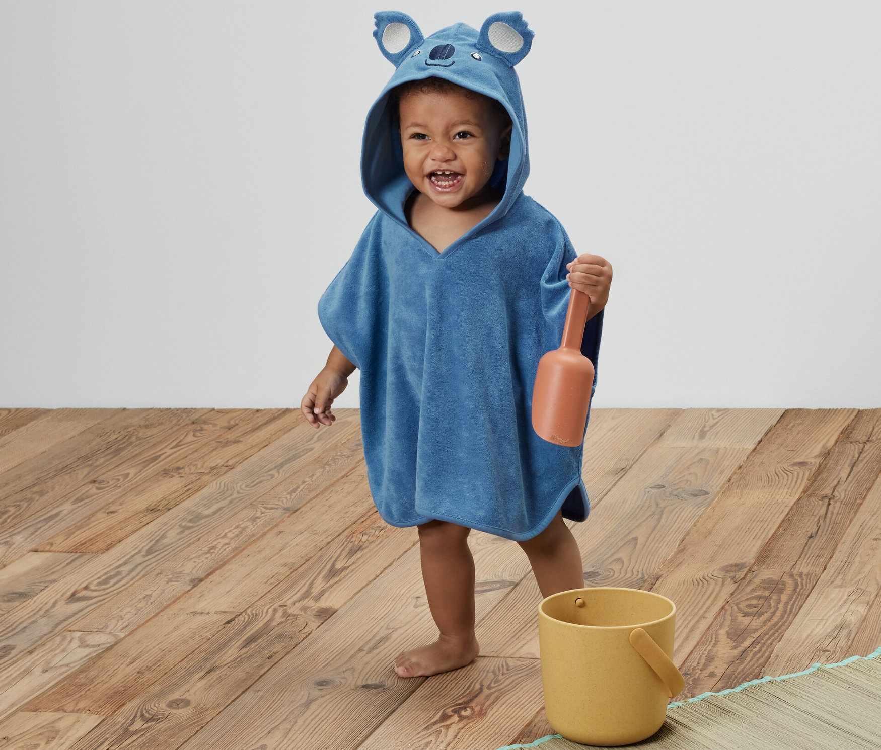 Un enfant portant un poncho en tissu éponge bleu avec une capuche en forme de koala tient une pelle orange. Un seau jaune est à côté de lui.