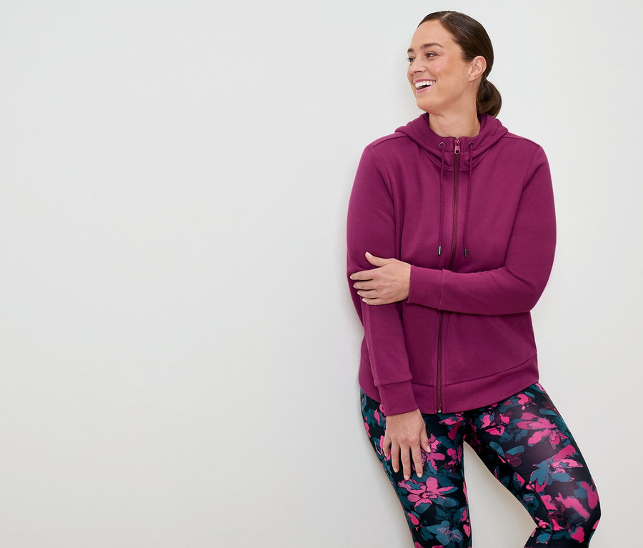Une femme portant un sweat à capuche zippé violet et un legging à fleurs, appuyée contre un mur blanc et souriant.
