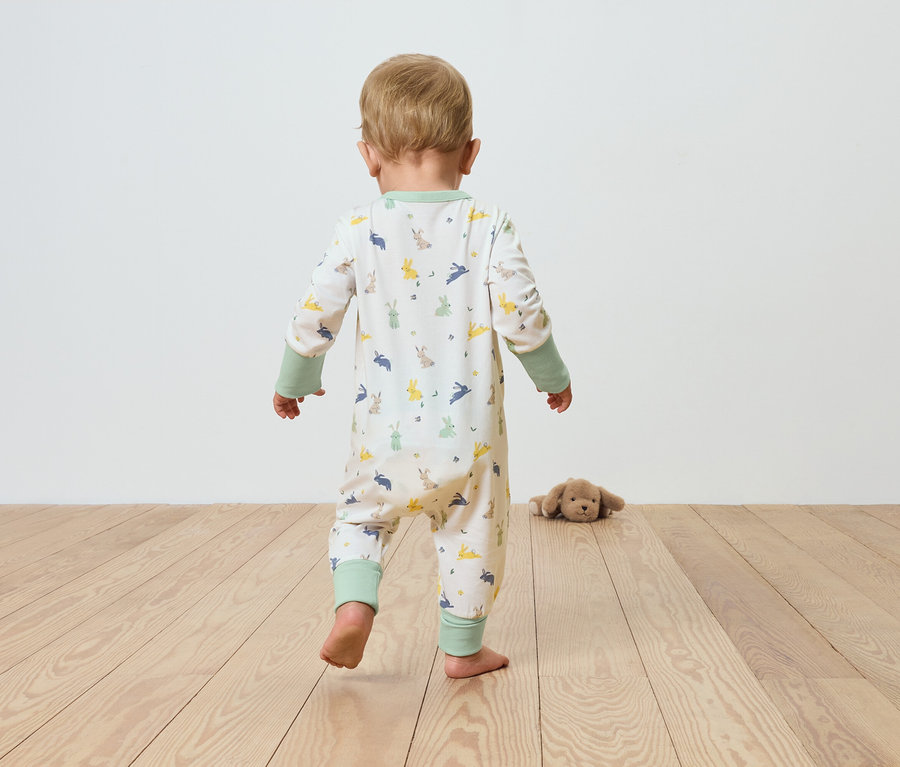 Un bébé en pyjama à motifs marche sur un plancher en bois en direction d'une peluche.