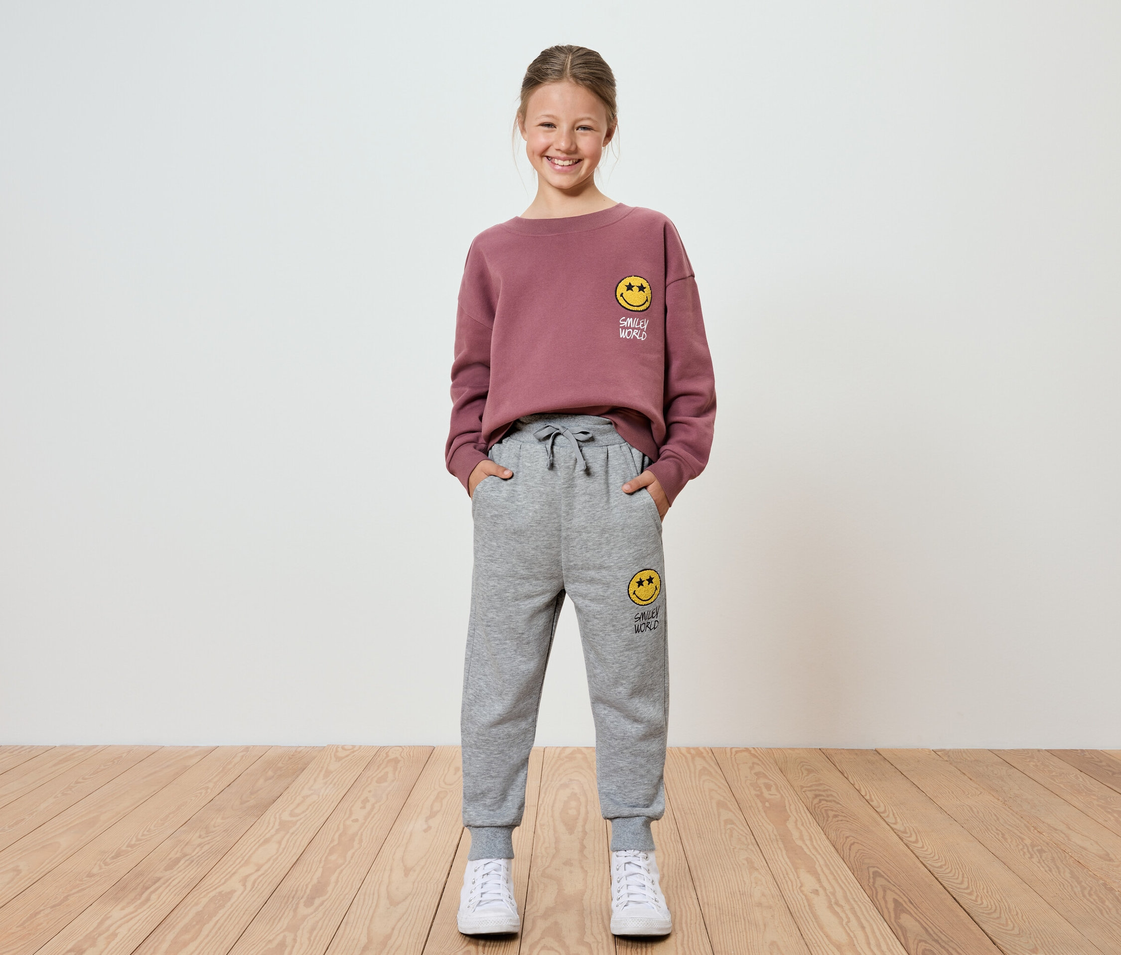 Une fille en sweatshirt bordeaux et pantalon de jogging gris avec un smiley se tient devant un mur blanc.