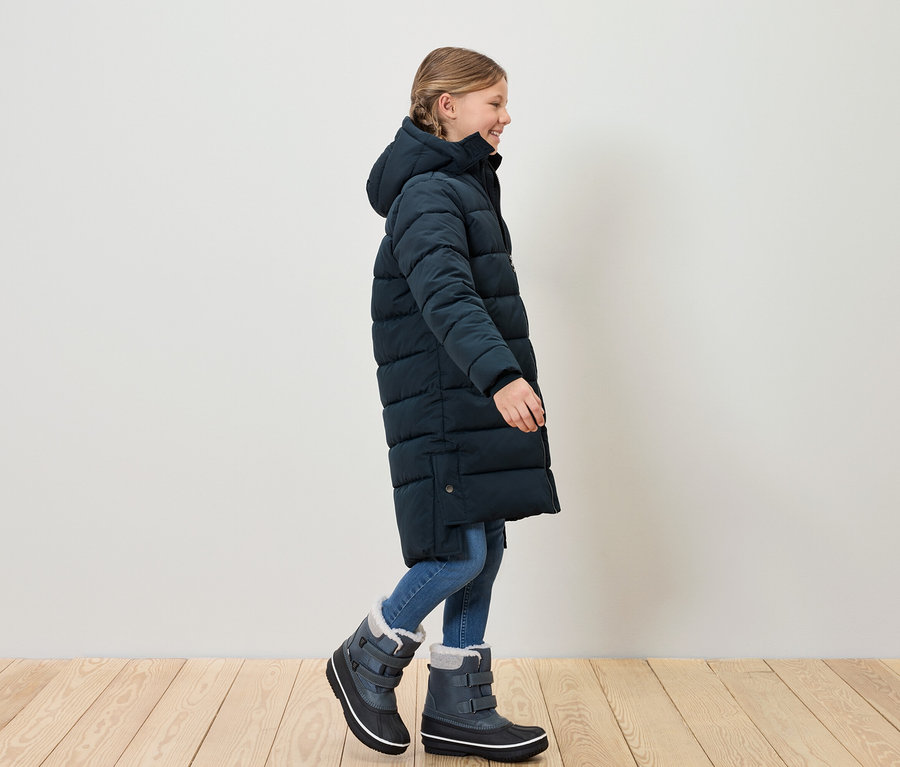 Une fille en manteau matelassé bleu et bottes de neige marche sur un plancher en bois.