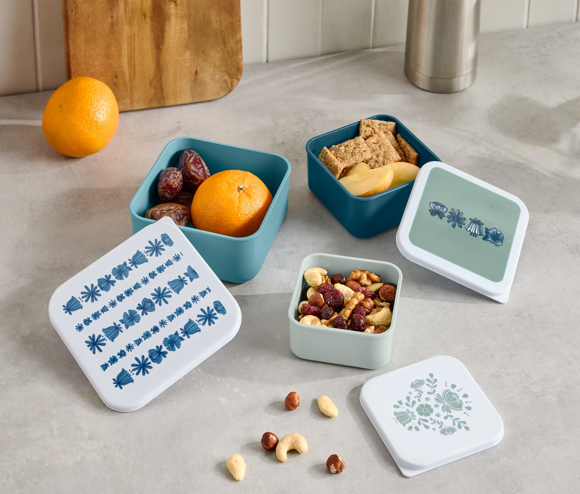 Sur un comptoir, une thermos et une planche à découper en bois sont posées à côté de quatre petites boîtes ouvertes contenant des fruits, des crackers et des noix, ainsi que quatre couvercles avec différents motifs.