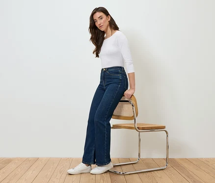 Une femme est assise de côté sur une chaise, portant un jean bleu et un t-shirt blanc.