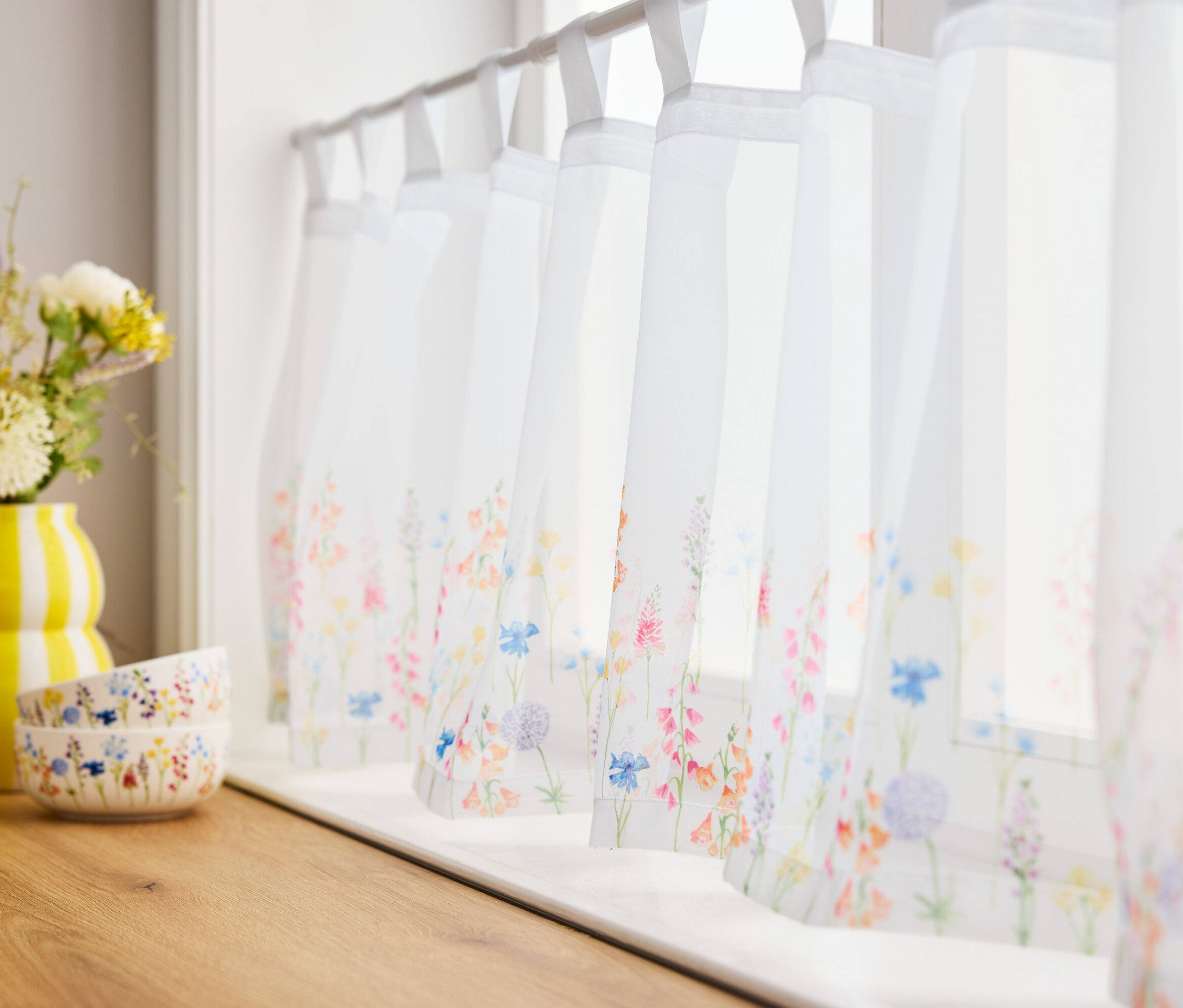 Fleurs dans un vase jaune et deux bols sur une planche en bois devant des fenêtres avec des rideaux à fleurs.