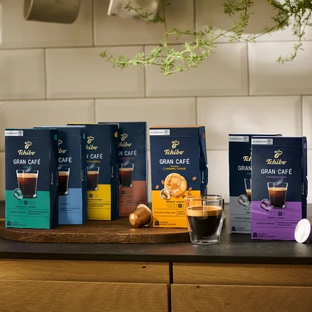 Sur un plateau en bois, des paquets de capsules de café Tchibo Gran Café de différentes saveurs et un verre de café.