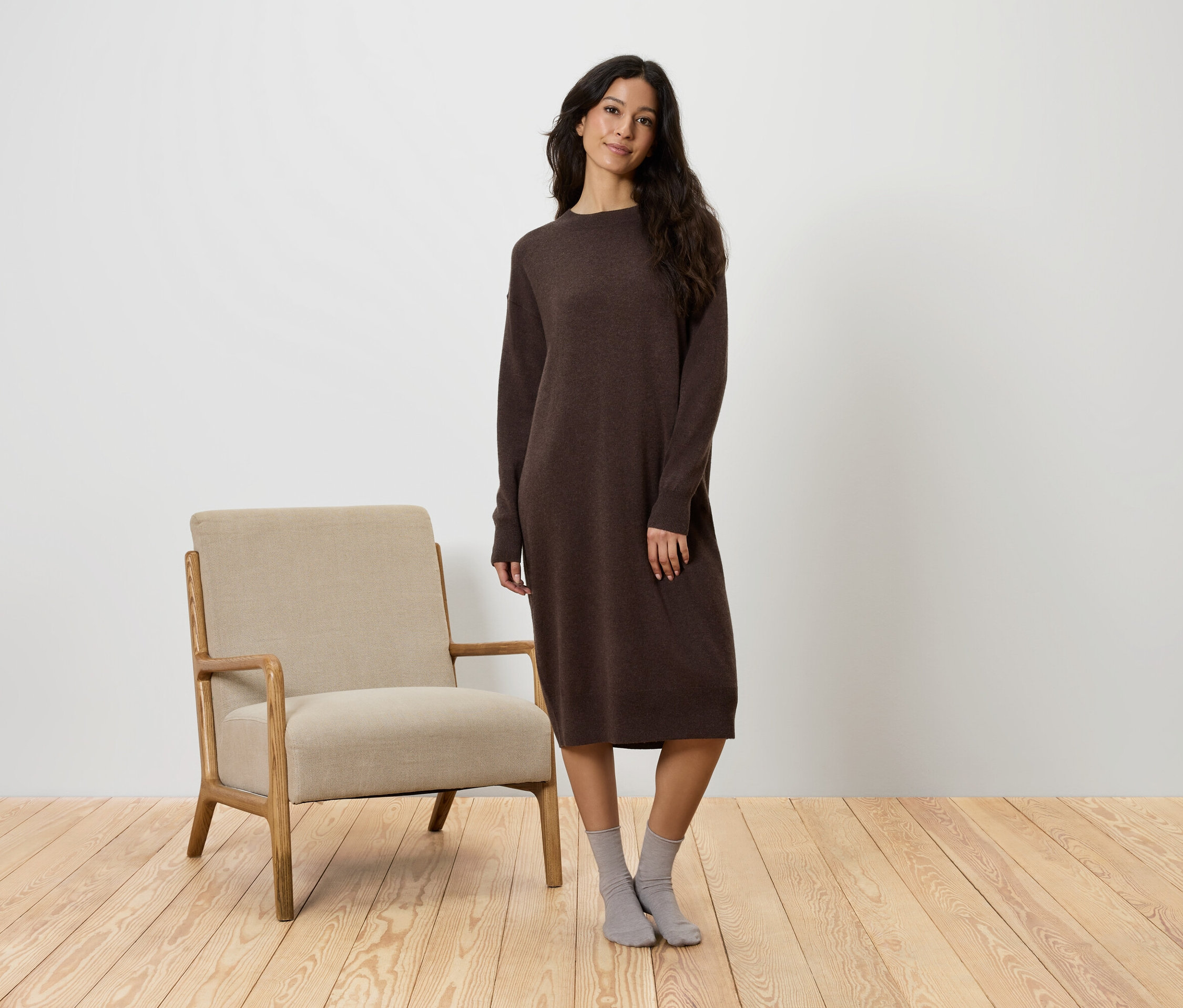 Une femme se tient à côté d'un fauteuil beige, portant une robe marron et des chaussettes grises.