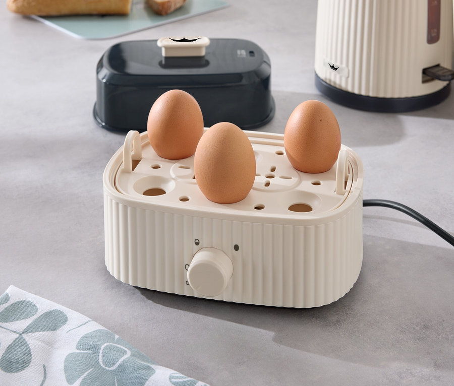 Trois œufs dans un cuiseur à œufs sur un comptoir de cuisine, avec un grille-pain, une bouilloire et du pain en arrière-plan.