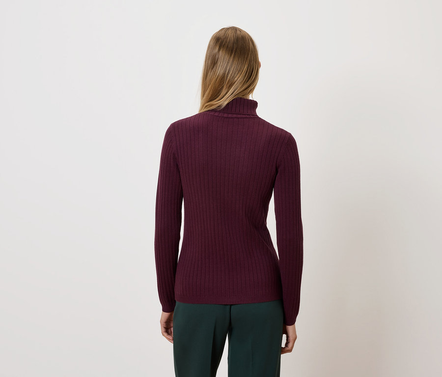 Une femme, vue de dos, porte un pull à col roulé bordeaux et un pantalon vert foncé.