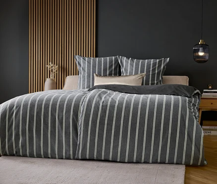Un lit avec des draps gris et blancs rayés devant un mur gris.