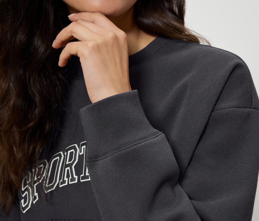 Gros plan sur une femme aux cheveux ondulés et foncés touchant son menton. Elle porte un sweat-shirt gris foncé avec l'inscription « Sport ». Gros plan sur la manche du sweat-shirt.