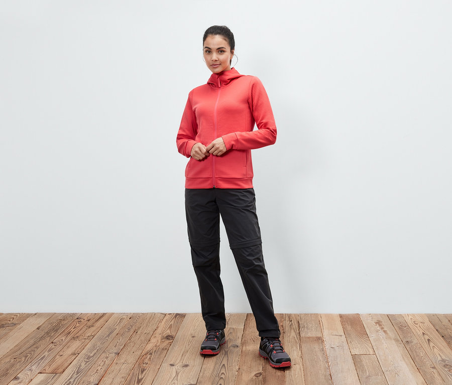 Une femme pose en veste à capuche corail, pantalon noir et chaussures sur un plancher en bois.
