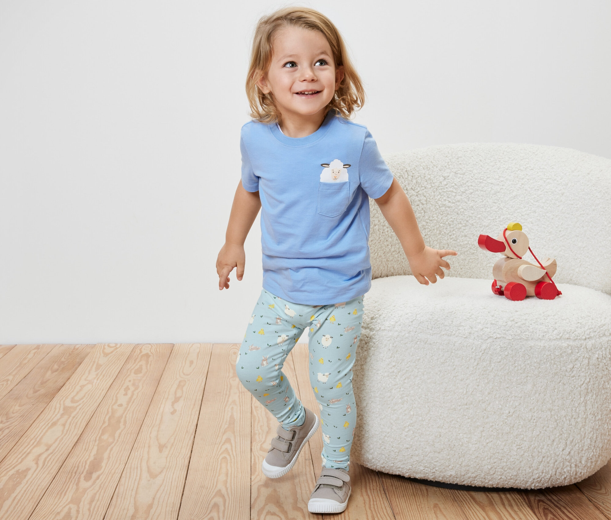 Un petit garçon portant un t-shirt bleu à manches courtes et 2 leggings pour enfant, bleu, se tient à côté d'un fauteuil blanc sur lequel est posé un jouet en bois.