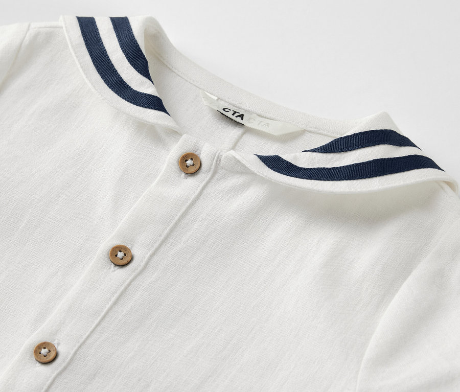 Gros plan d'une barboteuse de style marin blanche pour bébé avec des rayures bleu foncé sur le col et des boutons marron.