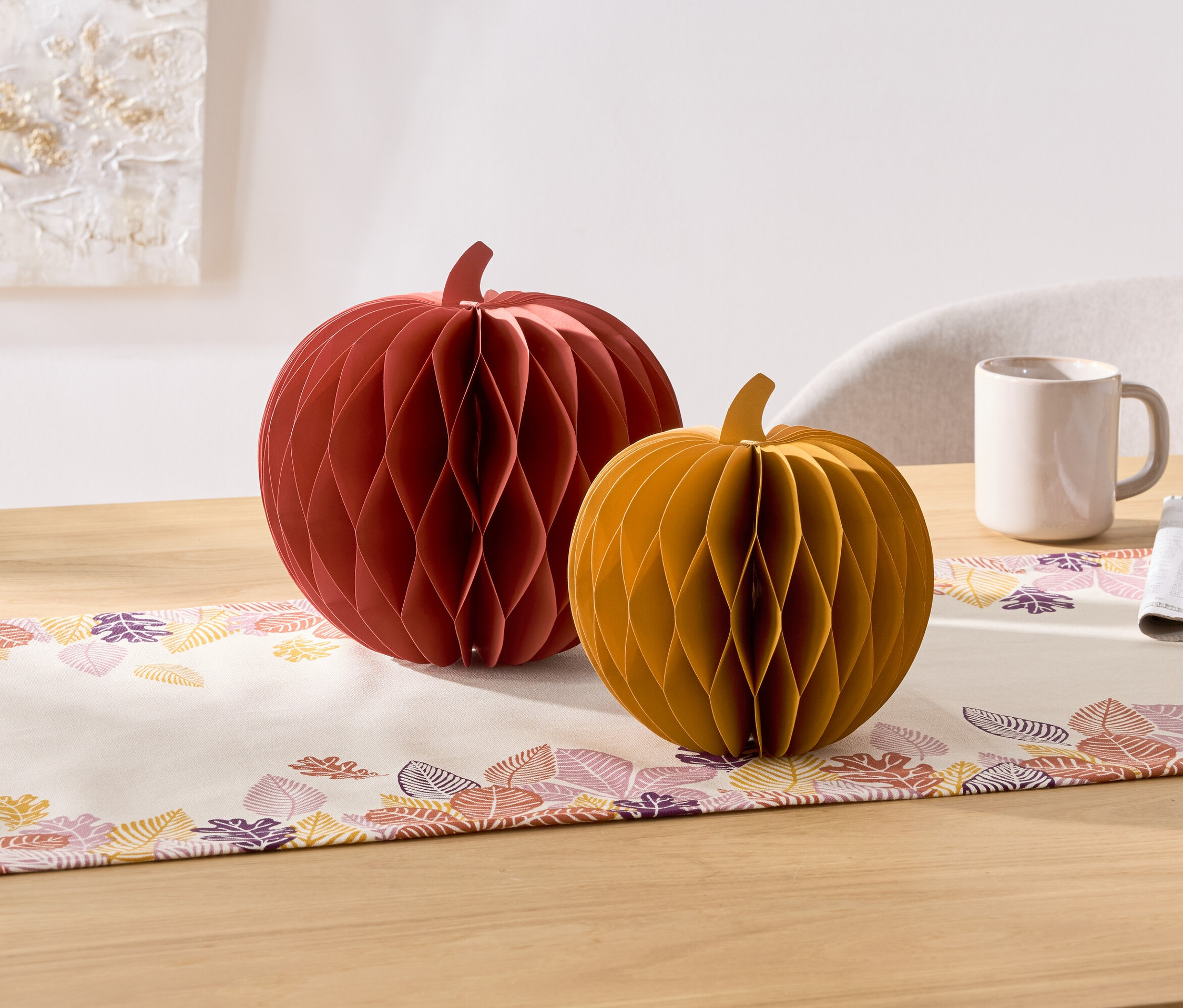 Sur une table, il y a deux citrouilles décoratives en papier, une rouge et une jaune. À côté, il y a une tasse et une serviette.