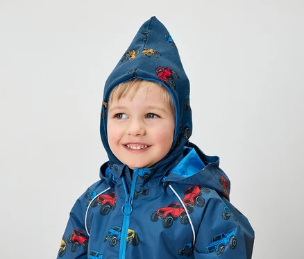 Un garçon souriant porte une veste bleue et une capuche avec un imprimé de monster trucks.