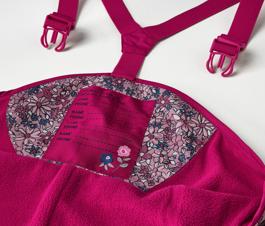Détail d'un pantalon de pluie thermique pour enfant, floral, incluant des détails comme les bretelles et l'étiquette de nom.