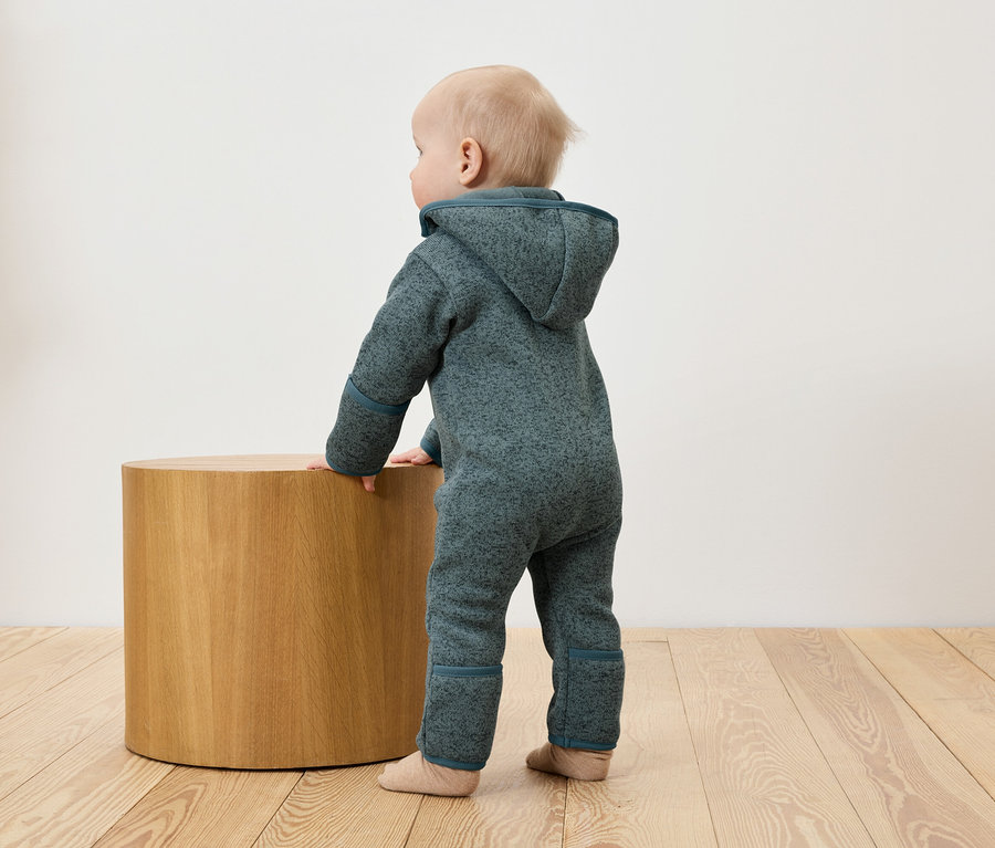 Un bébé en combinaison bleue s'appuie sur une table ronde en bois.