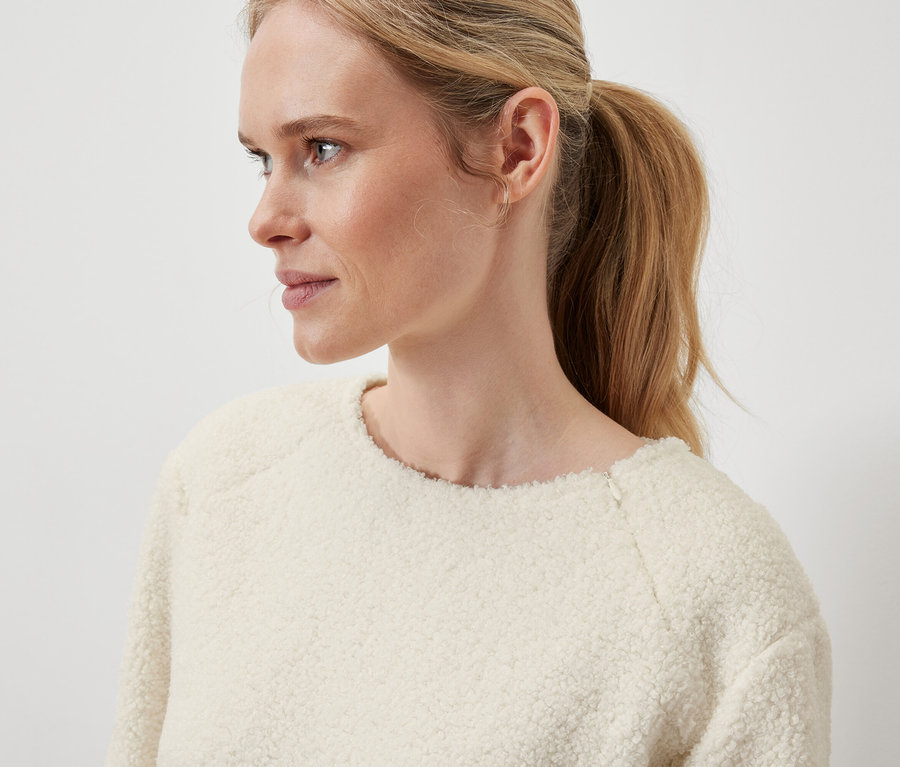 Gros plan sur le profil d'une femme blonde aux cheveux attachés en queue de cheval, portant un pull crème.