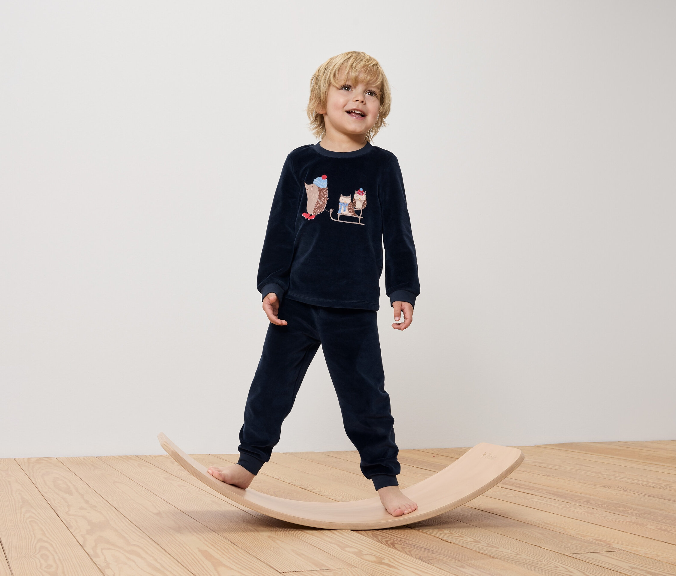 Un garçon en pyjama bleu marine avec des motifs de hérissons se tient debout sur une planche d'équilibre en bois.