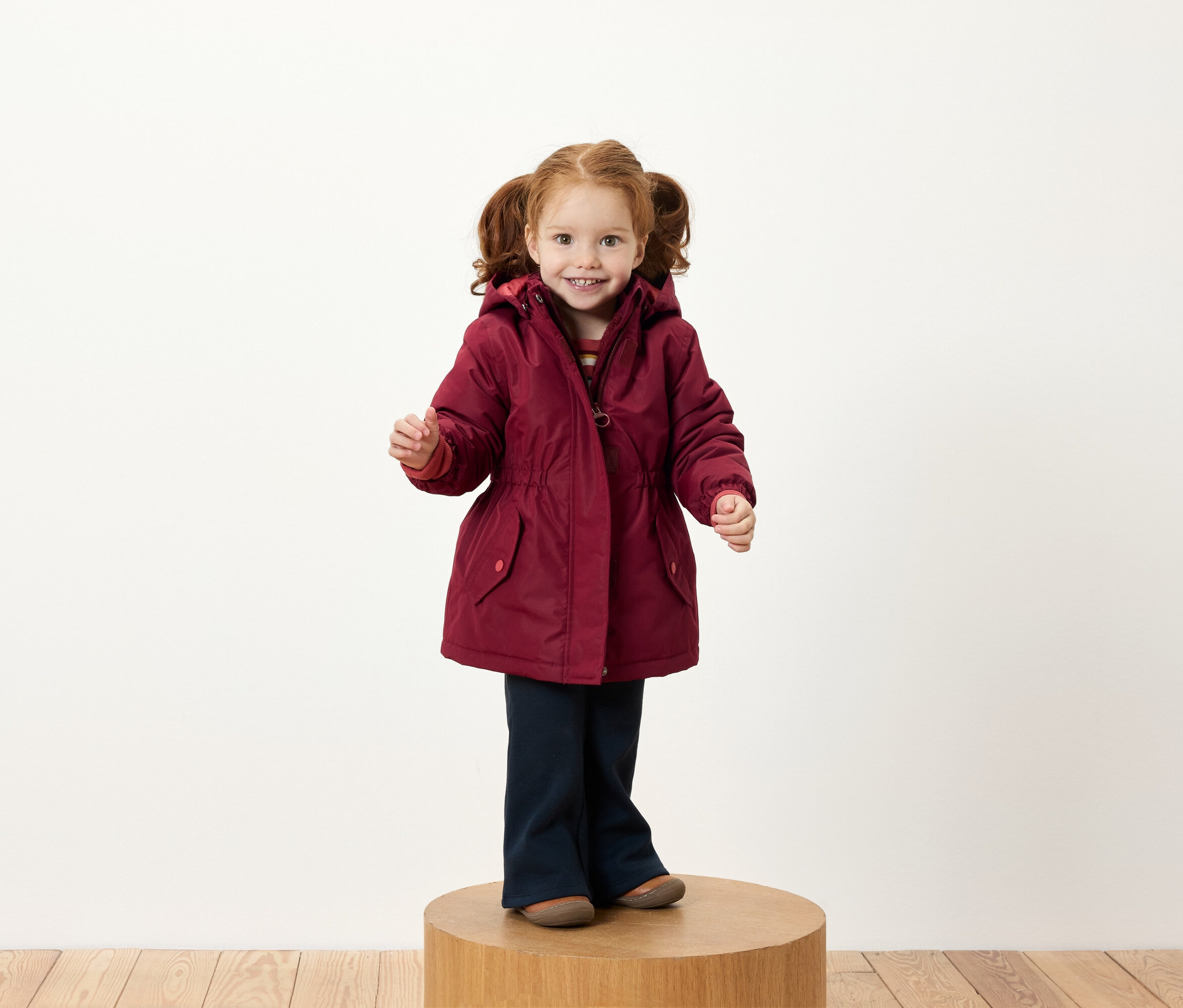 Une fille avec des couettes rousses se tient sur un socle en bois, portant une veste bordeaux et un pantalon bleu marine.