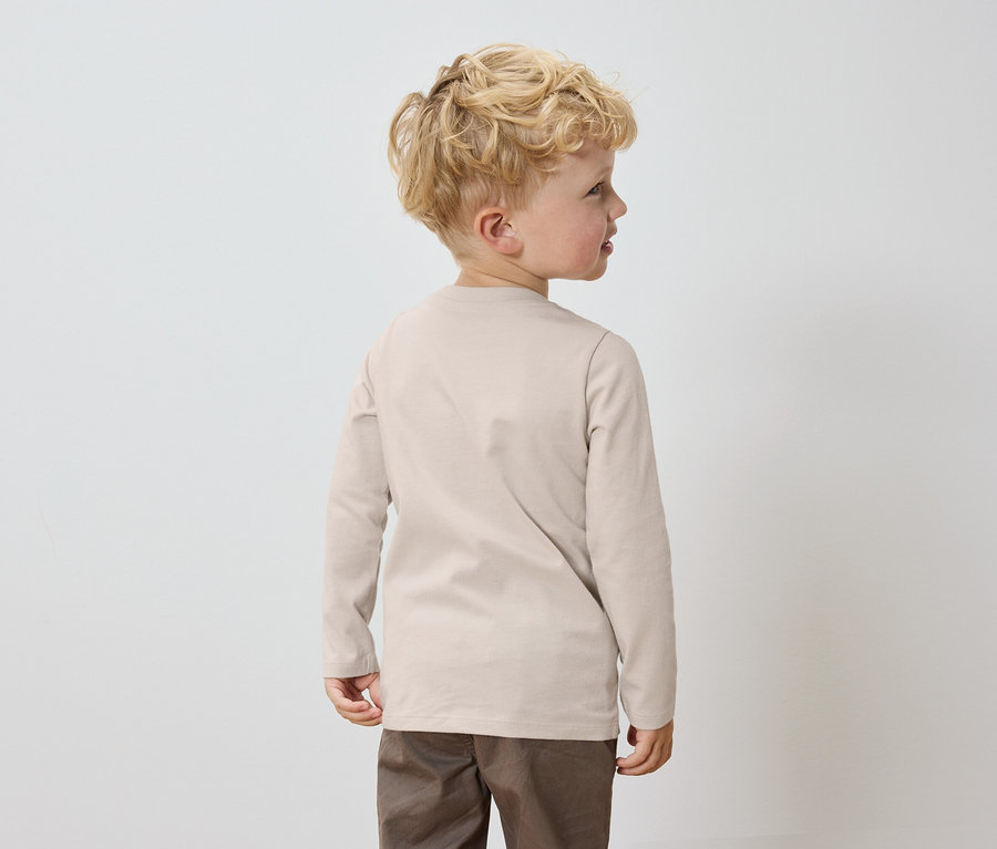 Un garçon aux cheveux blonds se tient dos à la caméra, portant un t-shirt beige à manches longues et un pantalon marron.