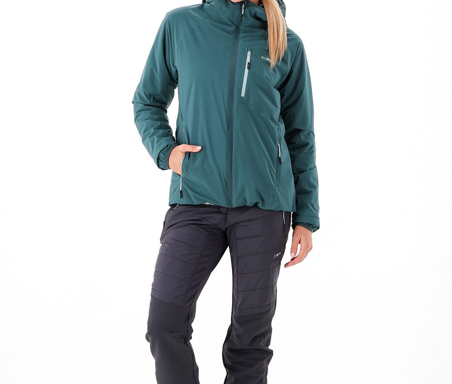 Une femme porte une veste vert foncé, un bonnet assorti et un pantalon de trekking hybride pour femmes CMP gris foncé avec des détails matelassés.