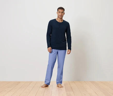 Un homme se tient pieds nus sur un plancher en bois, portant un t-shirt à manches longues bleu marine et un pantalon bleu clair.
