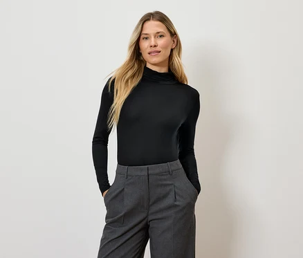 Femme aux longs cheveux blonds posant en pull à col roulé noir et pantalon gris.