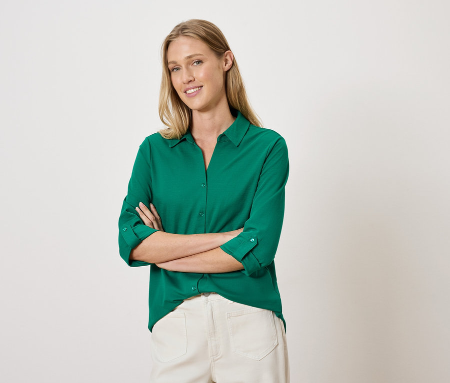 Une femme pose les bras croisés, vêtue d'une chemise verte et d'un pantalon blanc.
