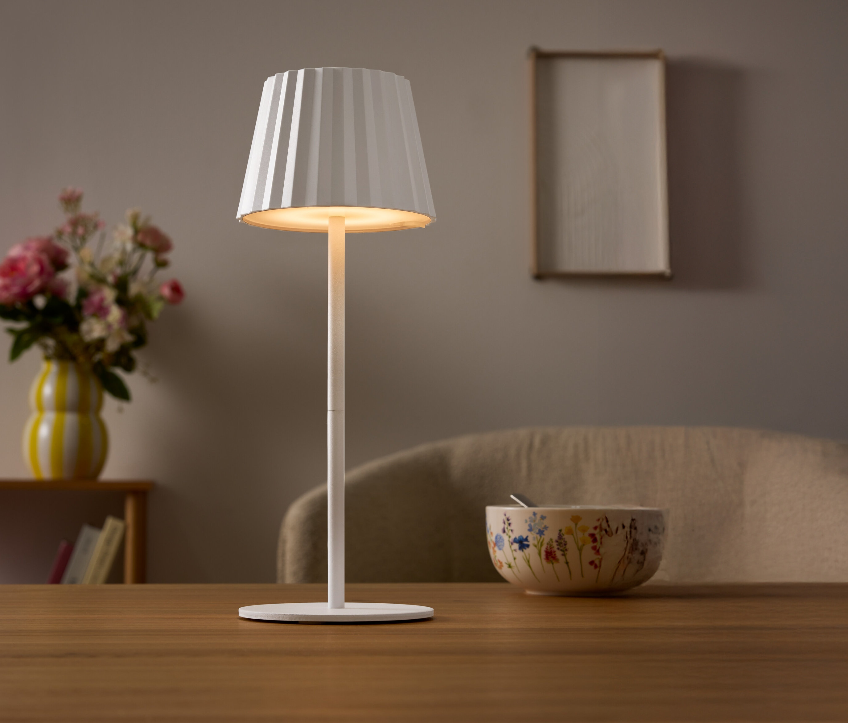 Une lampe blanche avec un abat-jour plissé est posée sur une table. Un vase de fleurs et un bol sont visibles à l'arrière-plan.
