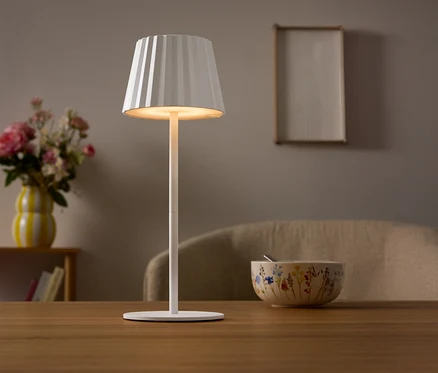 Une lampe blanche avec un abat-jour plissé est posée sur une table. Un vase de fleurs et un bol sont visibles à l'arrière-plan.