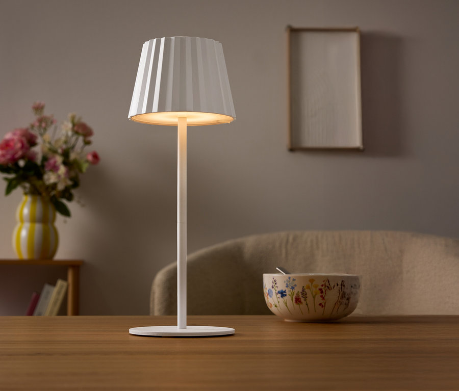 Une lampe blanche avec un abat-jour plissé est posée sur une table. Un vase de fleurs et un bol sont visibles à l'arrière-plan.