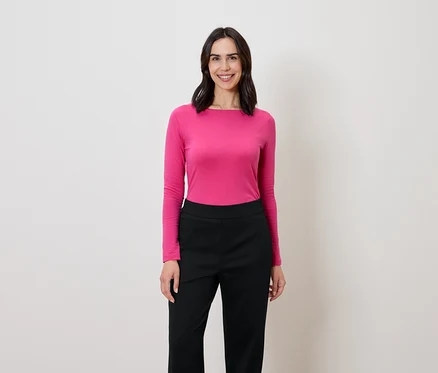 Une femme aux cheveux foncés se tient debout et sourit, elle porte un chemisier rose à manches longues et un pantalon noir.