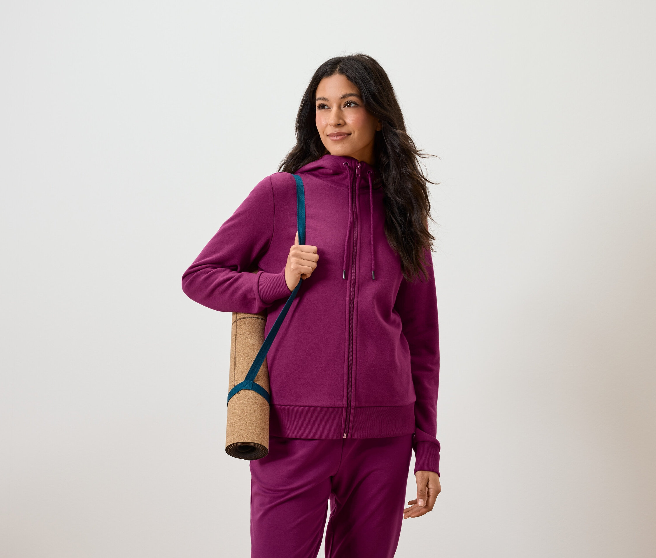 Femme aux cheveux foncés en survêtement violet tenant un tapis de yoga roulé sous le bras.